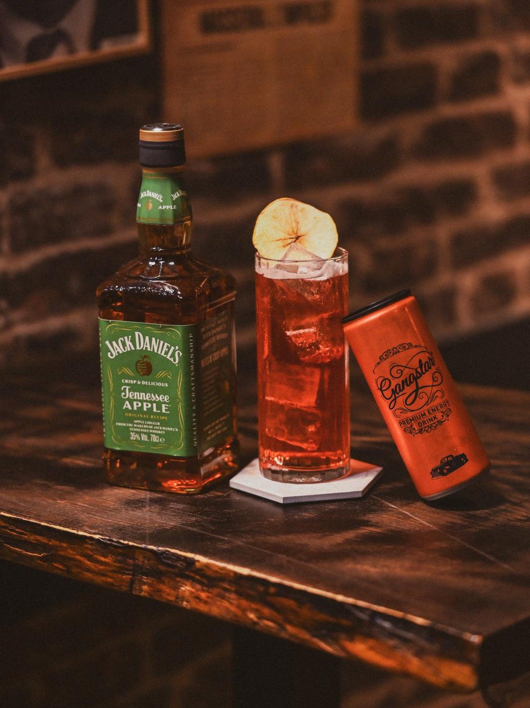 Le cocktail Jack Legs Diamond, orange et glacé, garni d'une pomme déshydratée, à côté d'une canette Gangstar