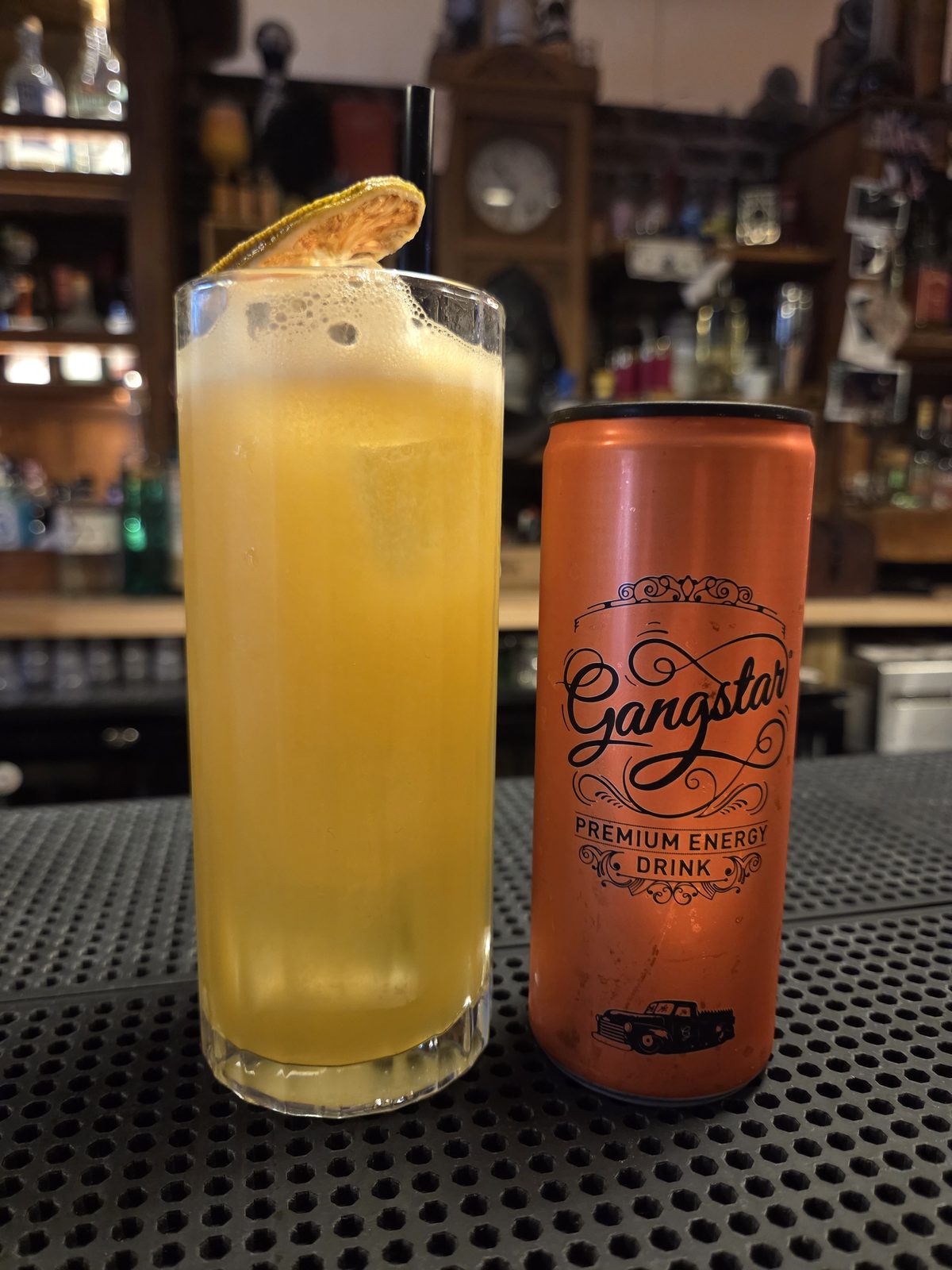 Le cocktail Golden Contraband, jaune doré dans un verre tumbler, garni d'une rondelle de citron déshydraté, à côté d'une canette Gangstar
