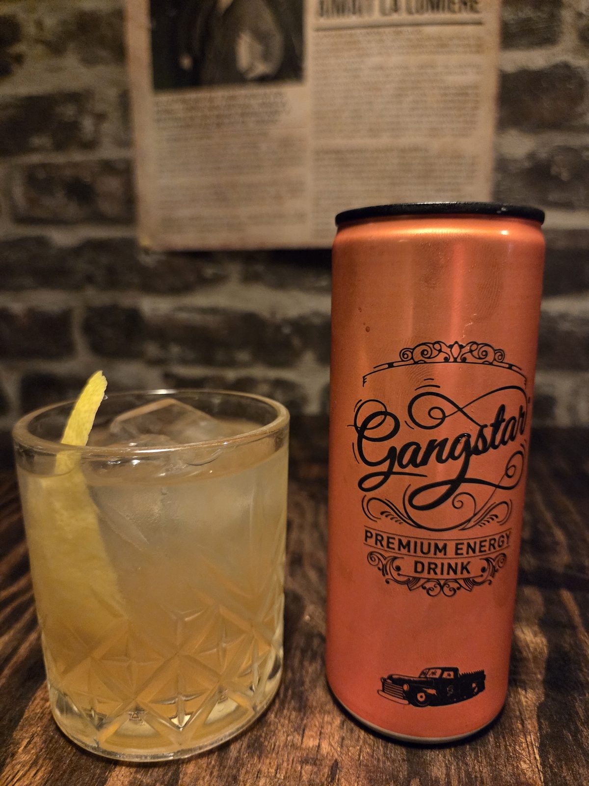 Le cocktail Copper Syndicate, doré dans un verre taillé, garni d'un zeste de citron jaune, à côté d'une canette Gangstar