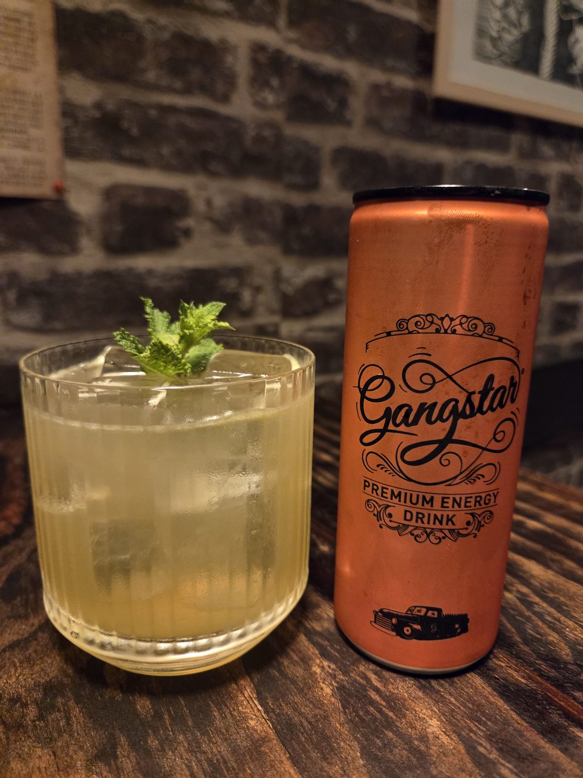 Le cocktail Rose Vendetta, dans un verre taillé, garni d'une tête de menthe, à côté d'une canette Gangstar